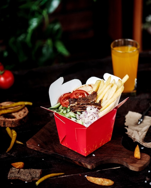 Side view of kebab meat with vegetable salad and french fries in cardboard bag on wooden cutting board