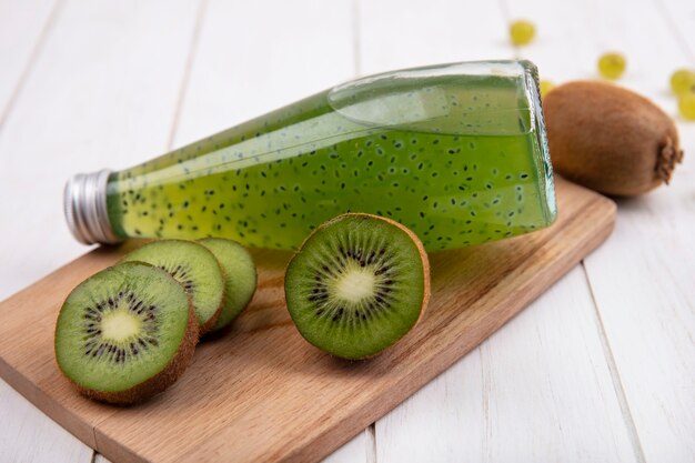 Side view kiwi wedges with a bottle of juice on a cutting board on a white wall