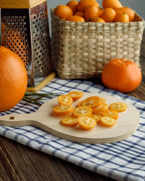 Vista laterale delle fette di kumquat sul tagliere con cesto di scalogno arancione mandarino di grattugia kumquat su panno a quadri su sfondo di legno