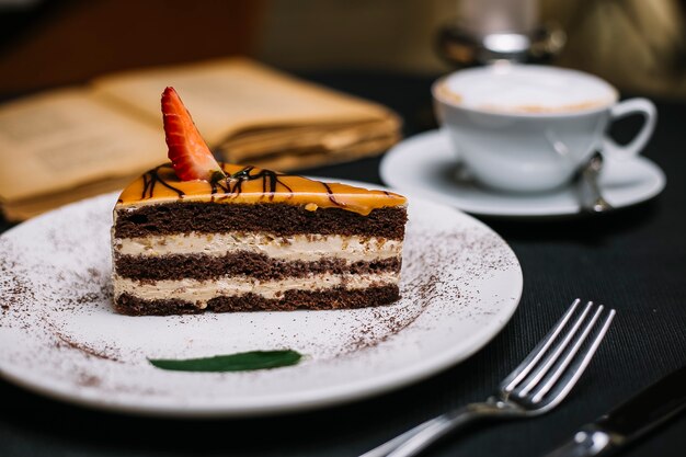 Side view of layered chocolate cake covered with caramel an strawberry slice on a white plate