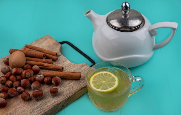 Side view of lemon juice with lemon slice in glass cup and nuts cinnamon walnut on cutting board with teapot on blue background