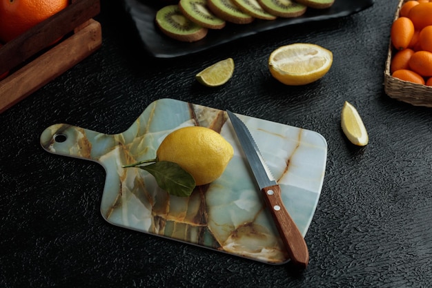 검은 배경에 키위 슬라이스 라임 슬라이스 컷 레몬과 전체 금귤이 있는 커팅 보드에 칼을 든 레몬의 측면 보기