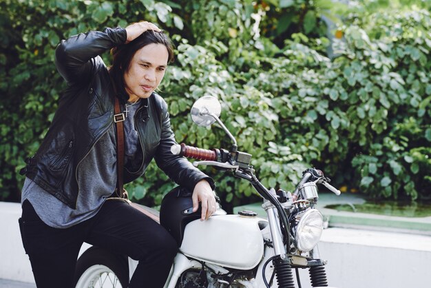 Side view of long-haired biker in leather jacket posing at his motorbike outdoors