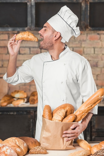 Side view of male baker eating fresh croissant holding loaf of breads