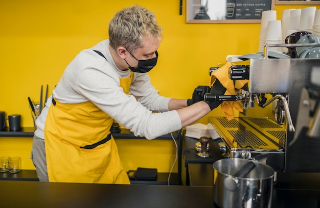 Foto gratuita vista laterale del barista maschio con macchina da caffè per la pulizia maschera medica