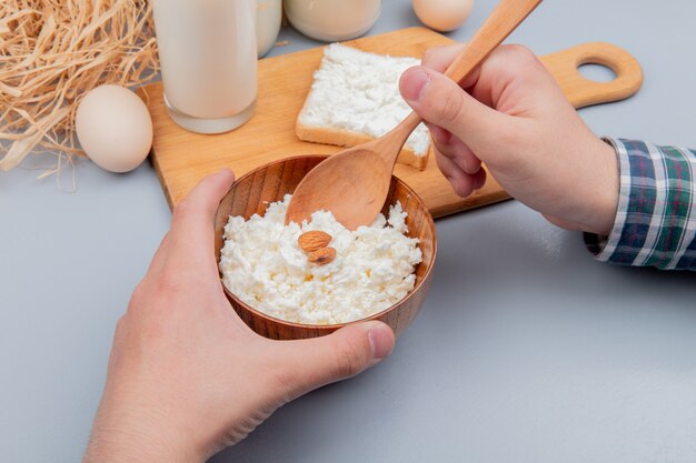 Side view of male hands holding bowl of cottage cheese and wooden spoon with bread slice smeared with cottage cheese milk on cutting board and eggs straw on blue surface