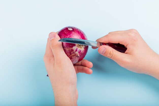 Side view of male hands peeling red onion with knife on blue background