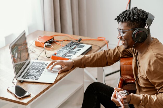 Мужской музыкант дома играет на гитаре и работает с ноутбуком, вид сбоку