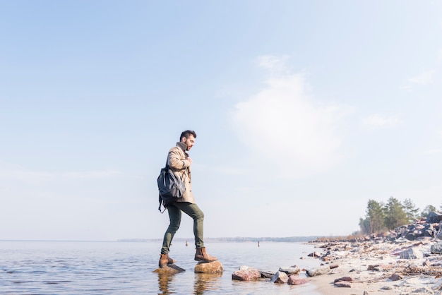 Вид сбоку мужского путешественника с его рюкзаком, стоя на камнях в озере