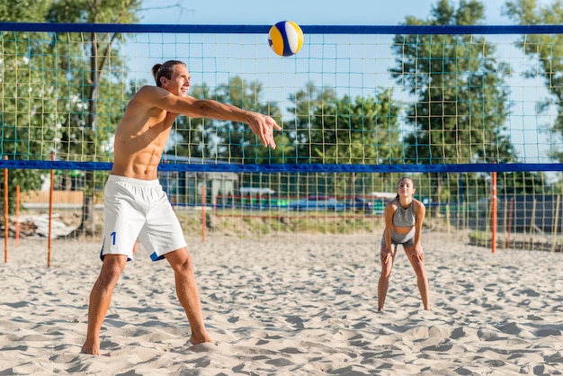 Вид сбоку мужского волейболиста на пляже с женщиной, играющей