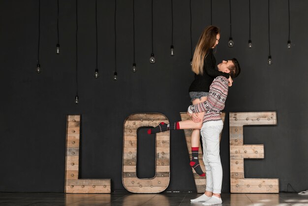 Side view of a man carrying her girlfriend standing in front of wooden love text against black background