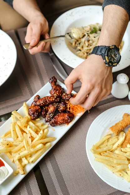 Side view man eating bbq chicken wings with fries and salad on the table