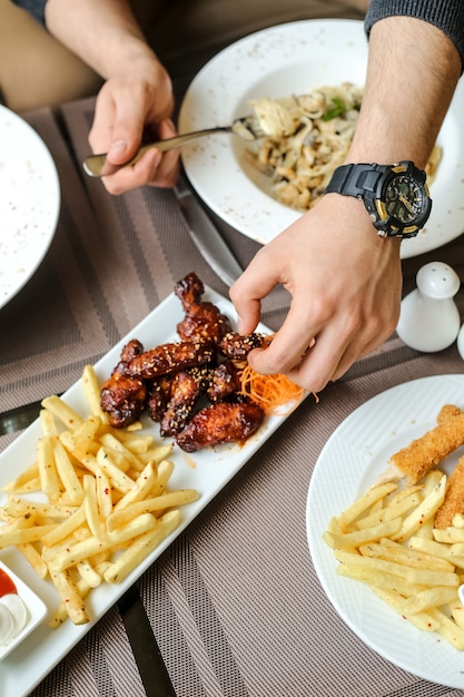 Side view man eating bbq chicken wings with fries and salad on the table