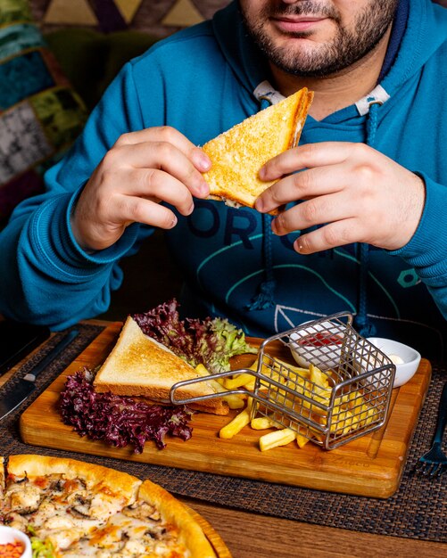 Side view of a man eating clubsandwich served with ketchup and french fries