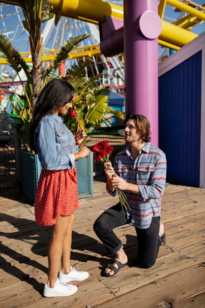 놀이 공원에서 무릎을 꿇고 여자 친구에게 장미 꽃다발을 제공하는 남자의 측면보기