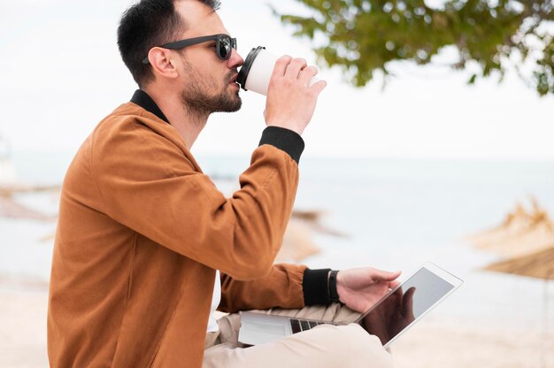 Side view of man having coffee at beach and working on laptop