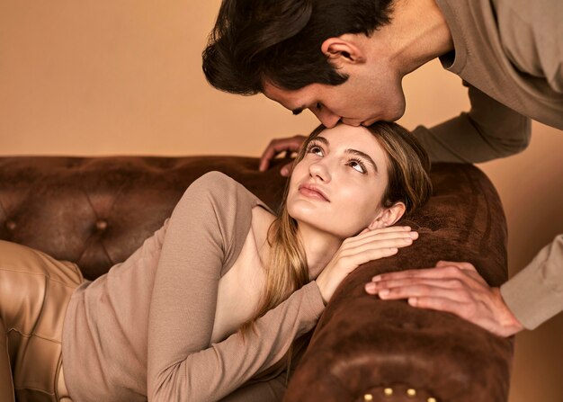 Side view of man kissing woman on the forehead while she sits on sofa