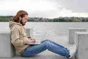 Free photo side view of man next to lake working on laptop