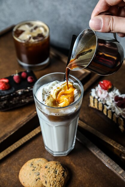 Side view a man pours coffee into milkshake with whipped cream in a glass with cookies on a tray