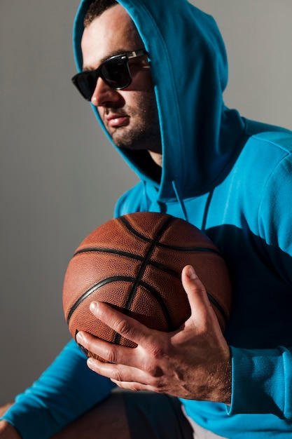 Side view of man with hoodie and sunglasses posing while holding basketball