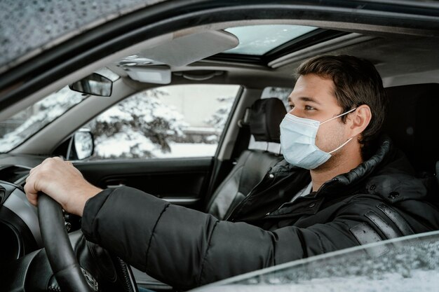도로 여행을 위해 차를 운전하는 의료 마스크를 가진 남자의 측면보기
