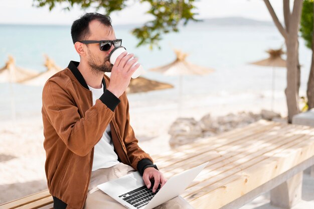 Side view of man with sunglasses having coffee at the beach and working on laptop