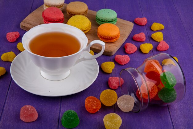 Side view of marmelads spilling out of jar and cup of tea on saucer with cookie sandwiches on cutting board and purple background