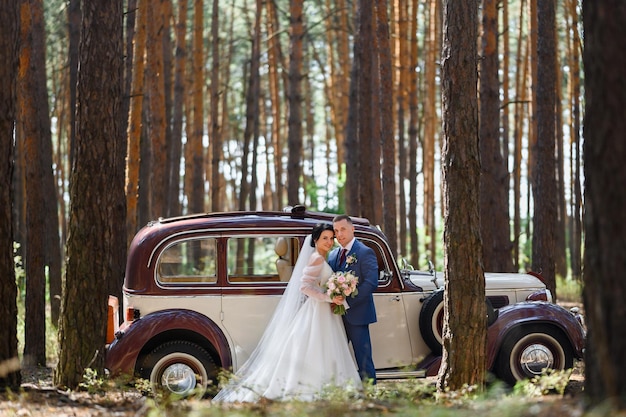 Side view of married bride and groom wearing in festive clothes standing face to face holding bridal bouquet smiling and looking at camera while posing near retro car in forest during wedding day
