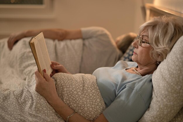 Side view of mature woman lying down in bed and reading book Her husband is sleeping in the background