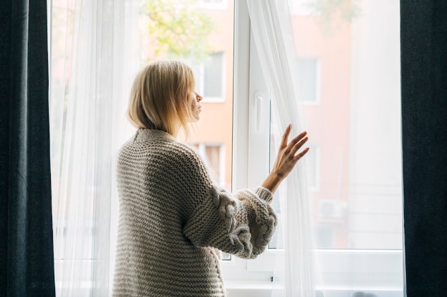 Вид сбоку меланхоличной женщины дома во время пандемии, смотрящей в окно