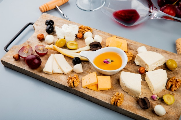 Side view of melted butter with different kinds of cheese grape olive nuts on cutting board and lying glass of wine on white