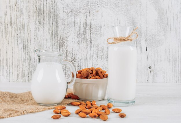 Side view milk carafe with bowl of almonds and bottle of milk on white wooden and piece of sack background. horizontal