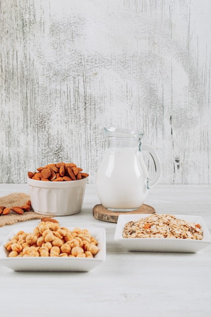 Side view milk carafe with bowl of almonds, hazelnut, and oats on white wooden background. vertical