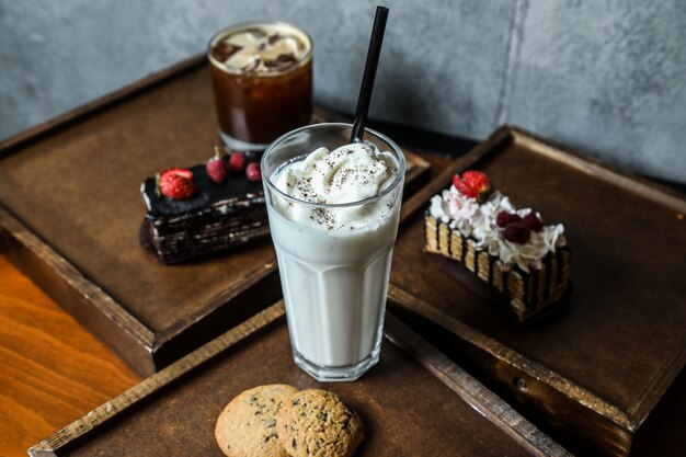 Side view milkshake with whipped cream and cookies on a tray with cakes