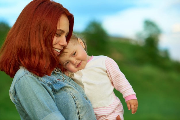 녹색 들판에서 포즈를 취하는 귀여운 어린 아이를 안고 있는 어머니의 옆모습 아름다운 얼굴을 가진 행복한 아이를 껴안고 있는 엄마는 가족 시간을 함께 보내는 청바지 셔츠를 입은 빨간 머리를 특징으로 합니다.