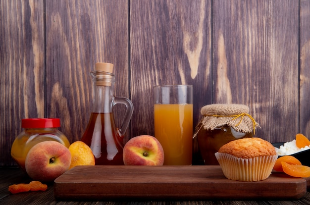 side view of a muffin on wooden board and fresh ripe peaches with a glass of peach juice and peach jam in a glass jar on rustic background