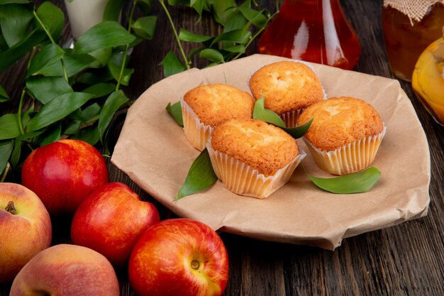 Side view of muffins with green leaves on craft brown paper with fresh ripe nectarines on rustic wood