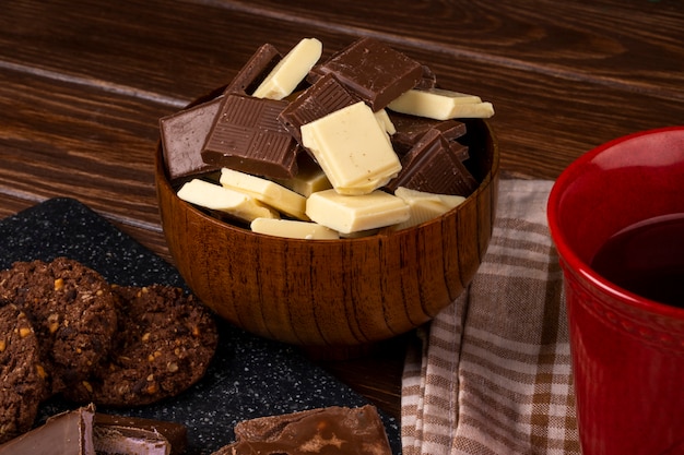 Foto gratuita vista laterale di una tazza con i biscotti di farina d'avena del tè e pezzi di cioccolato fondente e bianco in una ciotola di legno su fondo rustico