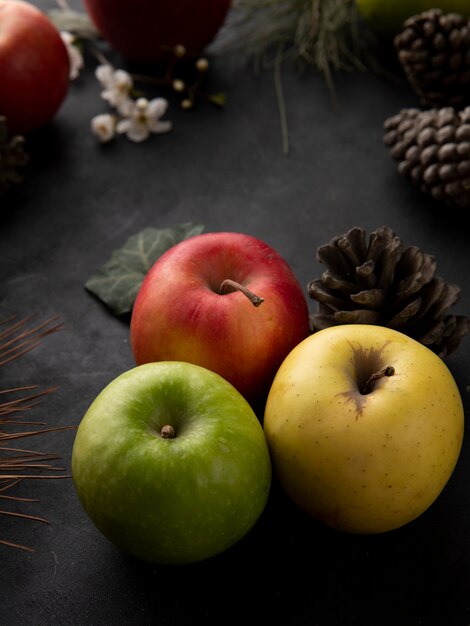 Вид сбоку разноцветных яблок с еловыми шишками на черной поверхности