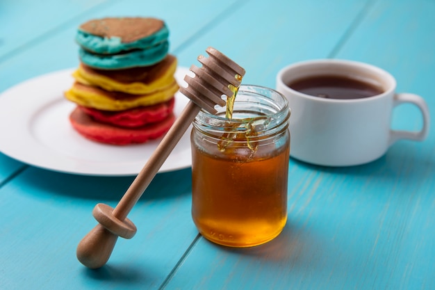 Side view multicolored pancakes on a plate with honey in a jar and a wooden honey spoon with a cup of tea on a turquoise background