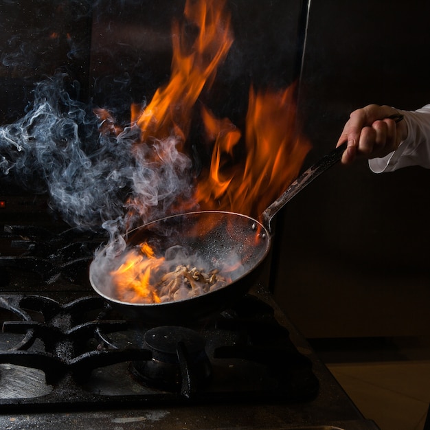 Side view mushroom frying with smoke and fire and human hand in pan