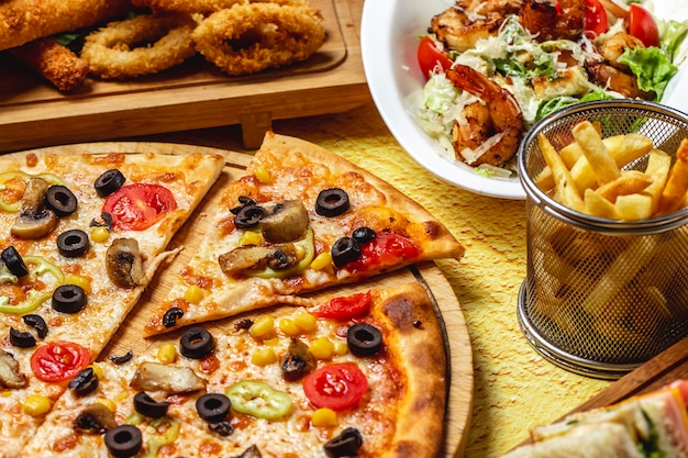 サイドビューマッシュルームピザとトマトコーンブラックオリーブチーズのフライドポテトとシーザーサラダテーブルの上のエビのグリル