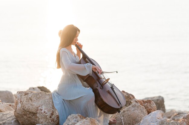 Вид сбоку музыканта, играющего на виолончели на скалах у моря