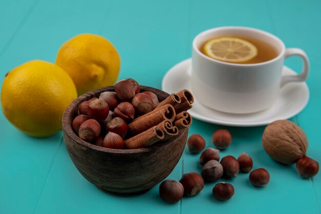 Side view of nuts and cinnamon in bowl with lemons and cup of tea on blue background