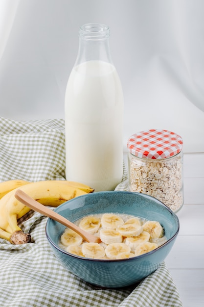 Side view of oatmeal porridge with banana in a ceramic bowl and a glass bottle of milk on rustic table