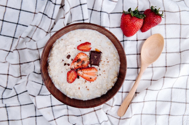 Side view of oatmeal porridge with fresh ripe strawberries and chocolate in a wood bowl and a spoon on plaid fabric