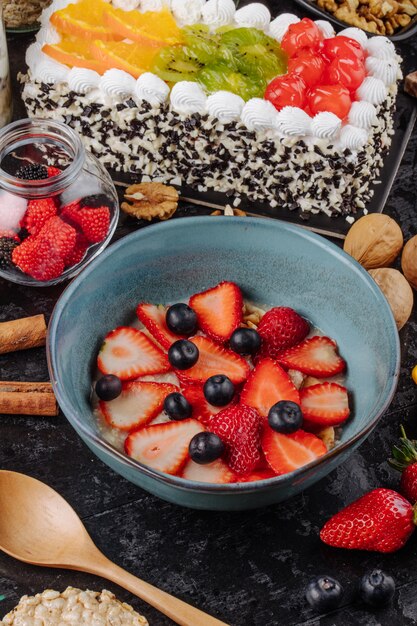 Side view of oatmeal porridge with sliced strawberries and blueberries in a ceramic bowl and fruit cake with whipped cream on the table