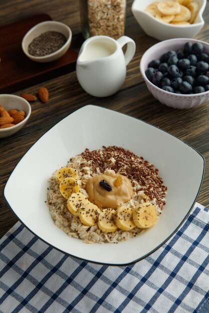 Side view of oatmeal with banana peanut butter sesame and dried fruits in plate on plaid cloth and milk blackthorn almond on wooden background