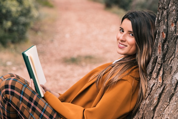 Бесплатное фото Взгляд со стороны усмехаясь молодой женщины сидя под деревом держа книгу в руке смотря камеру