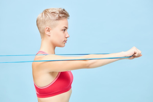 Бесплатное фото Вид сбоку атлетичной уверенно молодой женщины в красной верхней тренировке на синей стене с использованием полосы сопротивления, потянув ее, чтобы тренировать мышцы рук. концепция силы, энергии, решимости и мотивации
